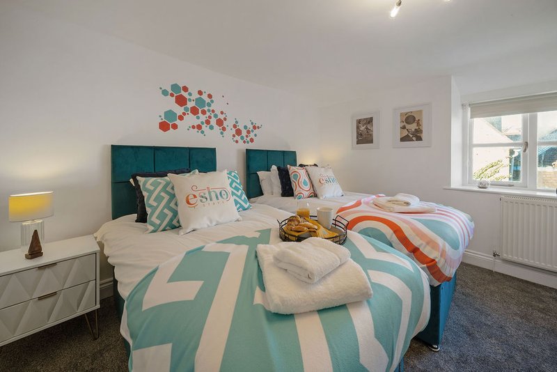 Bright bedroom with teal upholstered headboard, patterned bedding, wall art, and windows with radiator