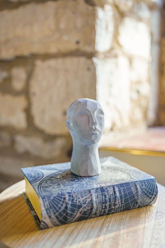 Decorative sculptural head on stacked books on wooden table, stone walls background