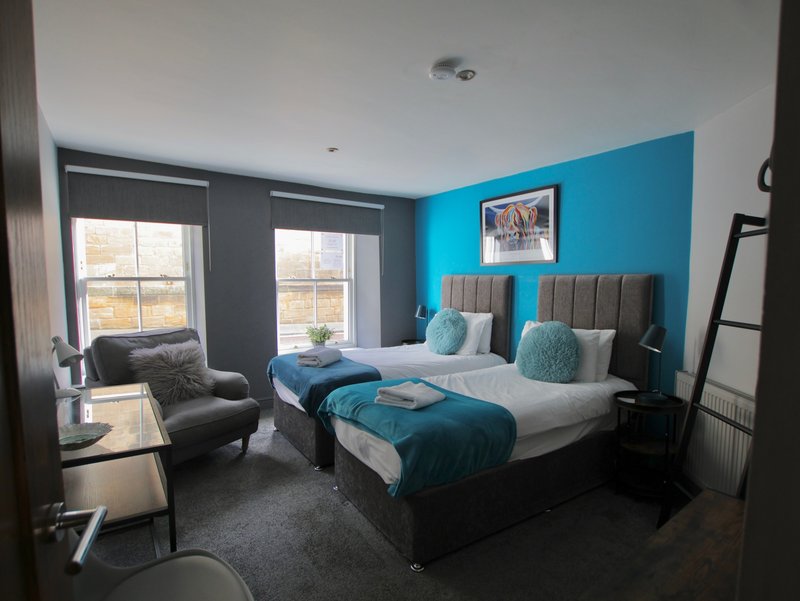 Modern bedroom with two single beds, turquoise accent wall, grey headboards and armchair by windows