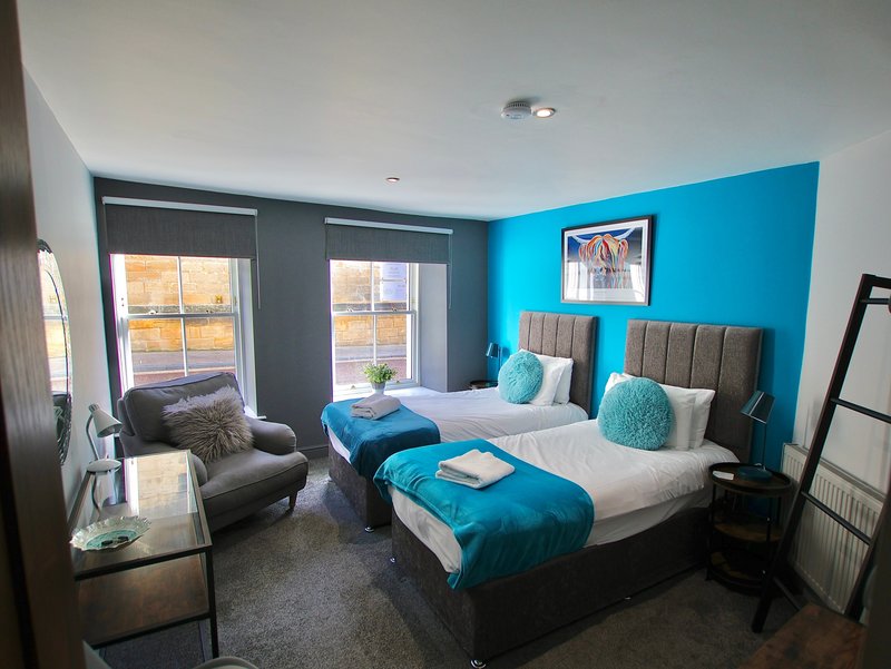 Modern bedroom with two single beds, bright blue accent wall, grey armchair and windows with city views