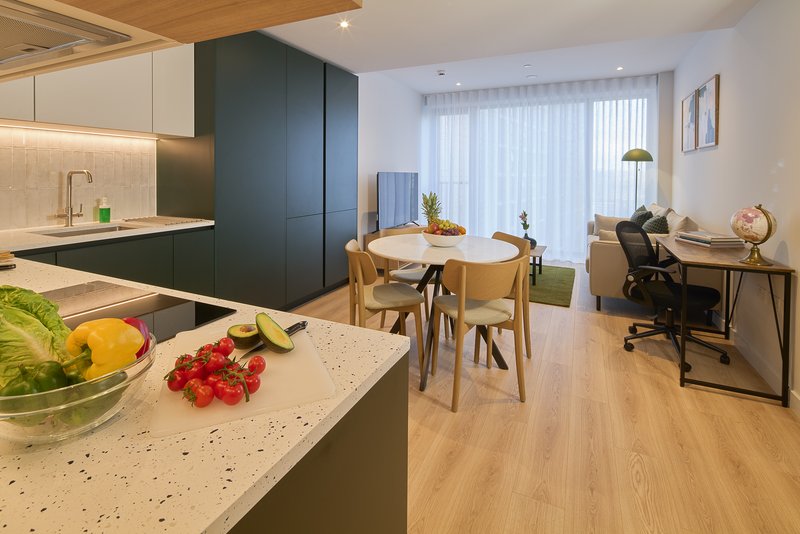 Open-plan kitchen and living area with dark cabinetry, wooden dining table, work desk, and fresh produce on counter
