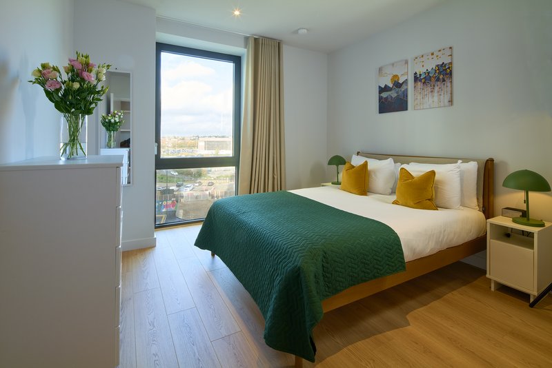Modern bedroom with double bed, green throw, wooden floor and city view from balcony