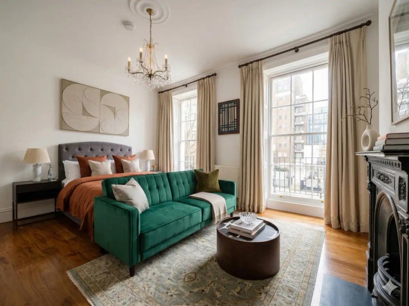 Bedroom with green velvet sofa, double bed with orange bedding, fireplace, and city views