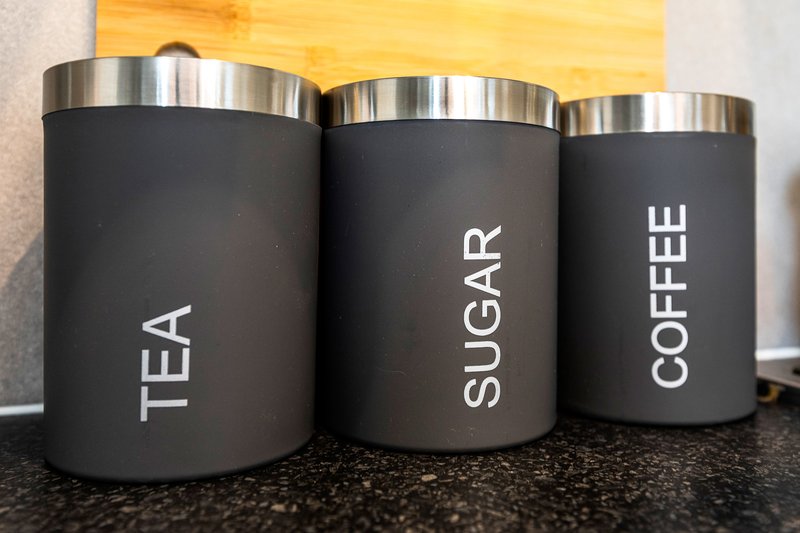 Three black kitchen storage containers with stainless steel lids labeled tea, sugar, and coffee