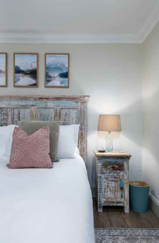 Bedroom with distressed wooden headboard, white bedding, pink cushions, framed landscape artwork, and bedside lamp
