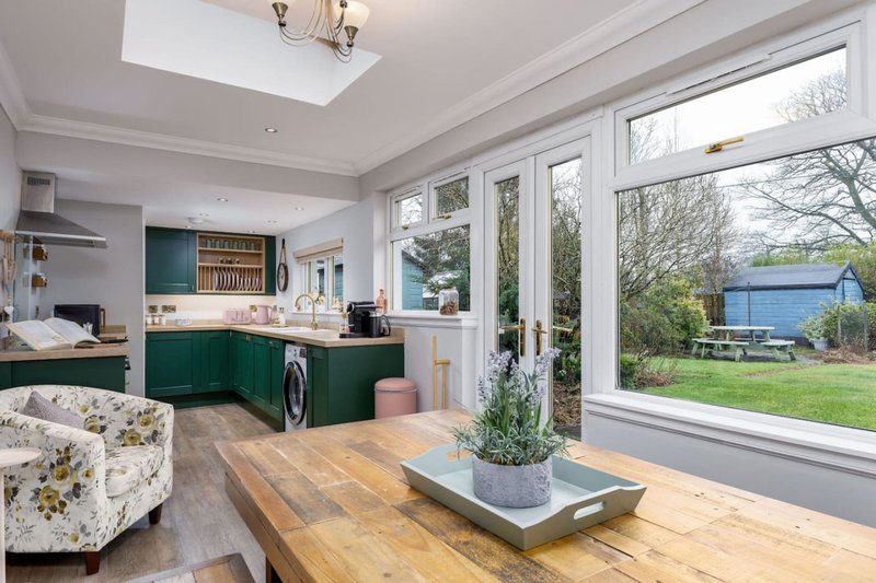 Open-plan kitchen and living area with green cabinetry, wooden table, and garden views through large windows