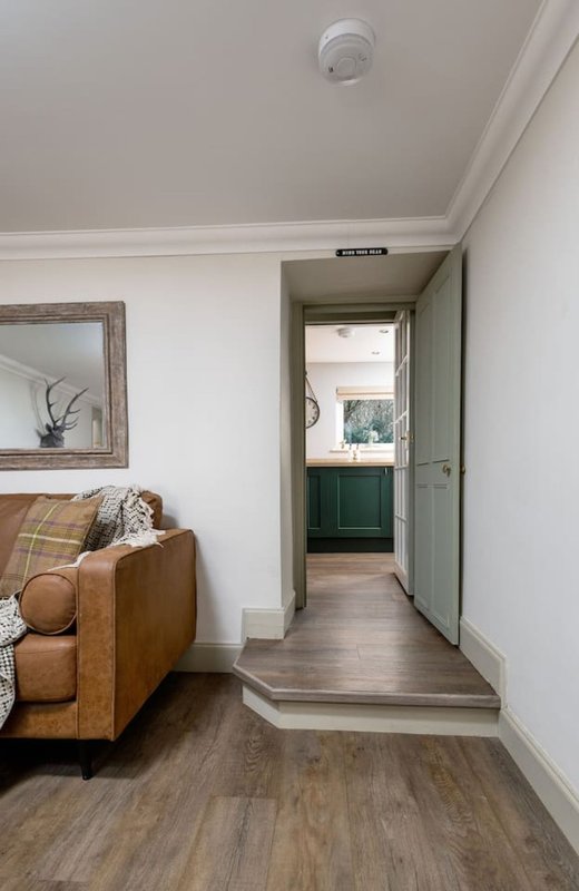 Living room with tan leather sofa, wooden flooring, white walls and open doorway to kitchen