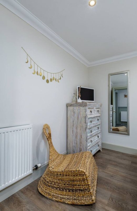 Bedroom with white walls, wooden dresser, small TV, woven chair, and gold decorative wall hanging