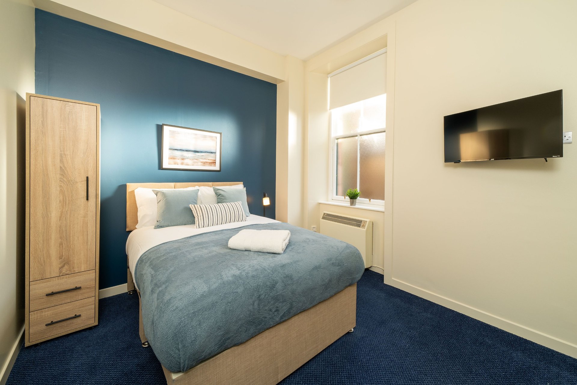 Modern bedroom with double bed, blue accent wall, wooden wardrobe, and wall-mounted TV