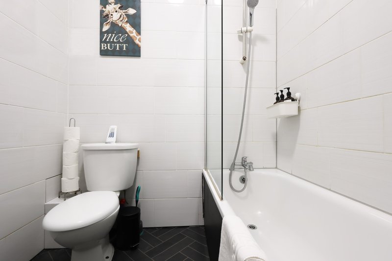 Small bathroom with white tile walls, toilet, bathtub with shower screen, and dark floor