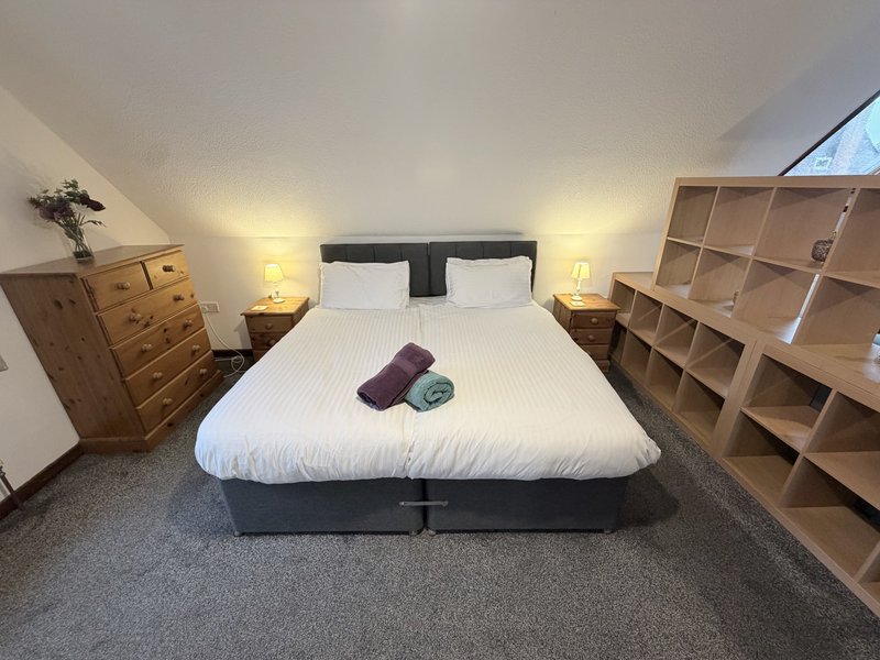 Bright bedroom with double bed, wooden chest of drawers, bedside lamps, and wooden shelving units