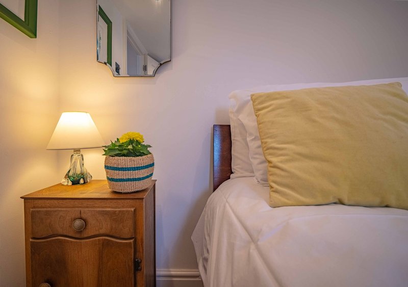Bedroom with wooden bedside table, lamp, mirror, and bed with yellow cushion