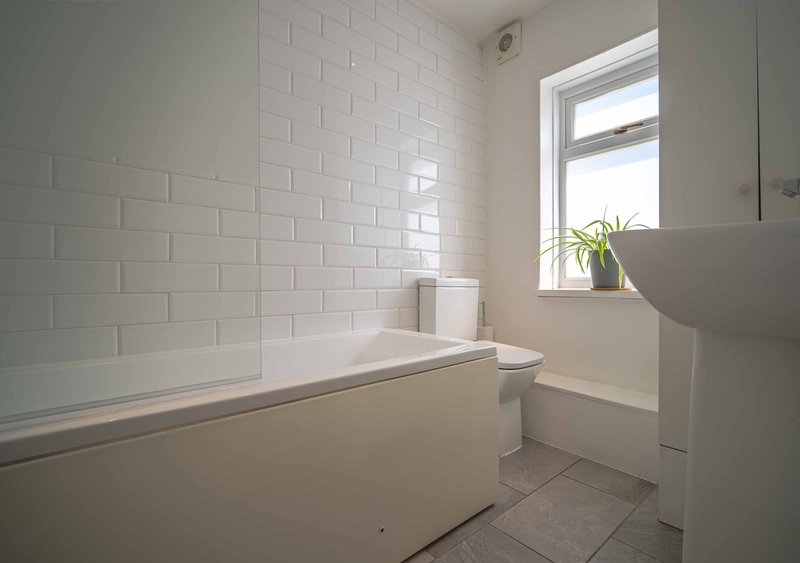 Clean bathroom with white subway tile walls, bathtub, toilet, and window with plant
