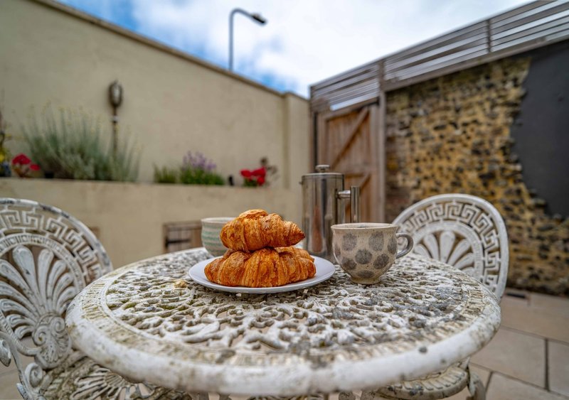 Outdoor patio table with croissants and cup, decorative chairs, stone walls and plants