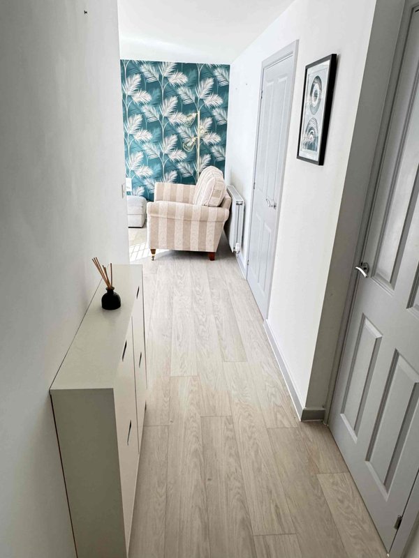 Bright hallway with light wood flooring, white walls, storage console, and seating area visible ahead