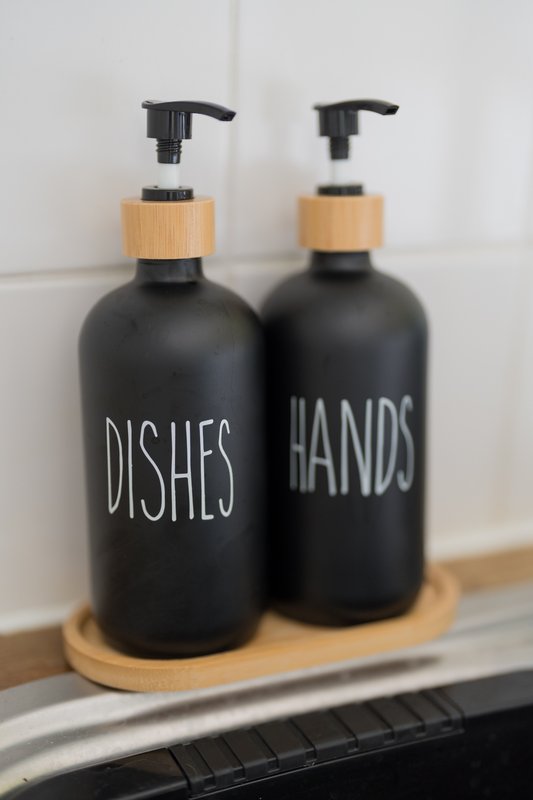 Two black soap dispensers with wooden tops labeled dishes and hands on kitchen counter