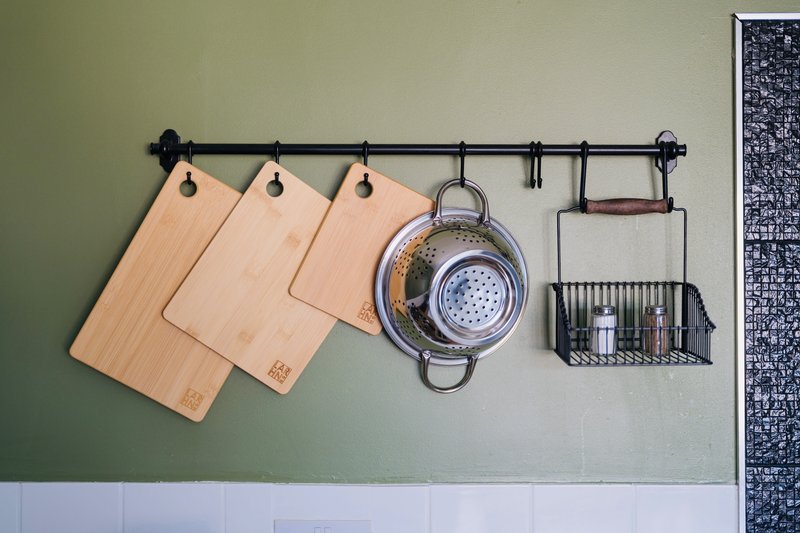 Kitchen wall rack with wooden cutting boards, metal colander, and storage baskets hung on black rail