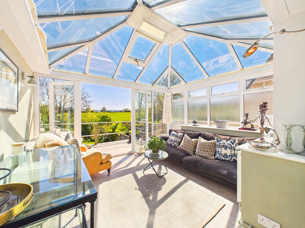 Bright conservatory with glass roof, dark sofa, yellow chair, and scenic countryside views