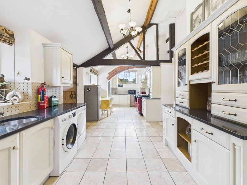 Galley kitchen with exposed beams, white cabinetry, black countertops, appliances and tiled floor.