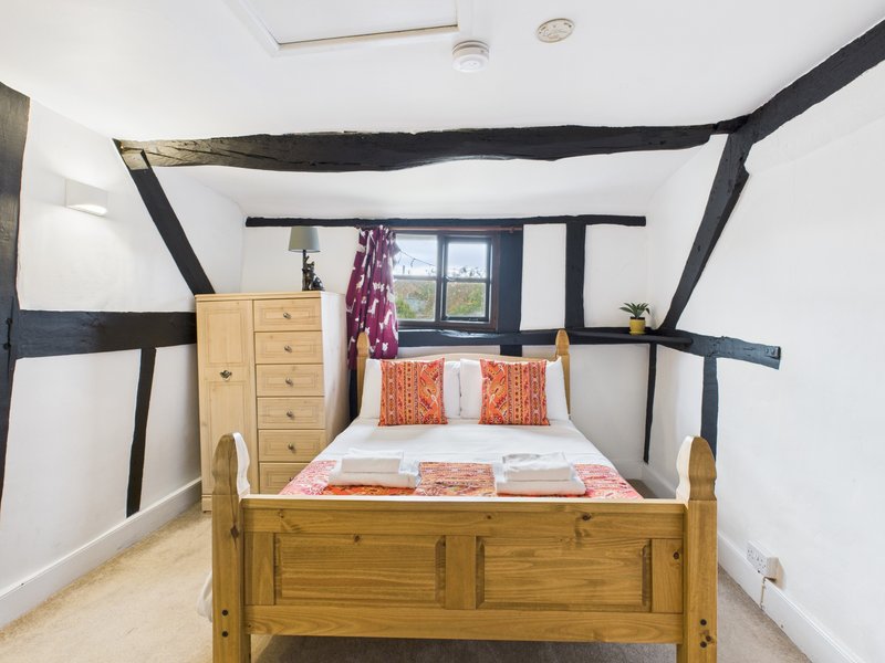 Cozy bedroom with wooden bed frame, orange cushions, exposed black beams, and window with countryside view