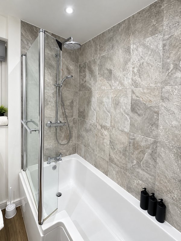 Modern bathroom with white bathtub, glass shower enclosure, grey tile walls and overhead rainfall showerhead