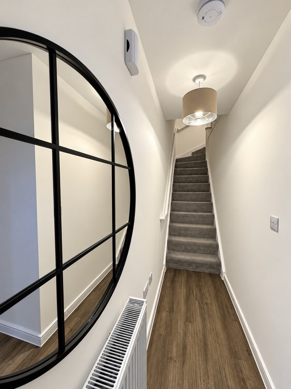 Narrow hallway with wooden flooring, white walls, stairs ahead, and black-framed mirror on left