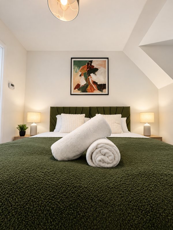 Modern bedroom with green upholstered headboard, white pillows, rolled towels, pendant light and framed artwork