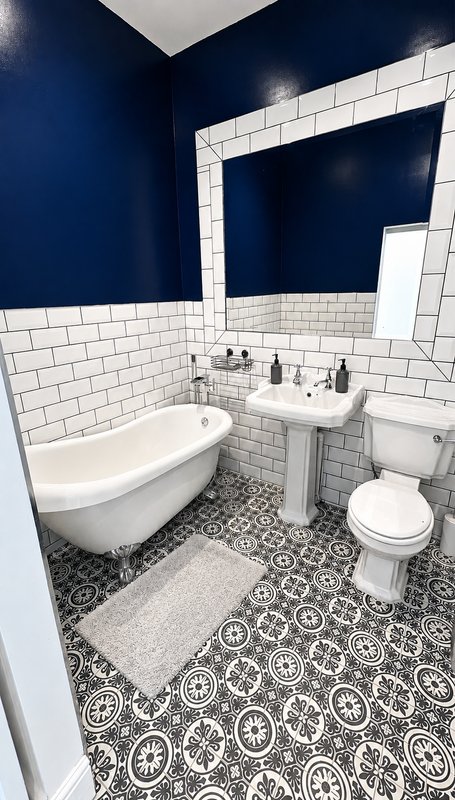 Modern bathroom with freestanding white bathtub, pedestal sink, toilet, navy walls and patterned tile floor
