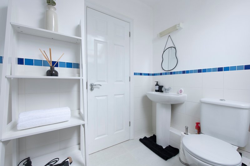 Clean white bathroom with pedestal sink, toilet, blue tile trim, and white shelving unit