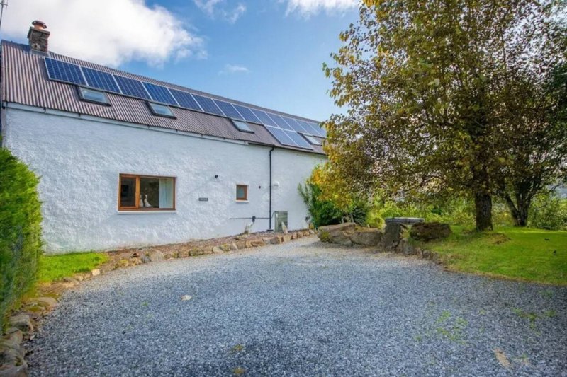 White cottage with solar panels, gravel driveway, and mature trees surrounding property