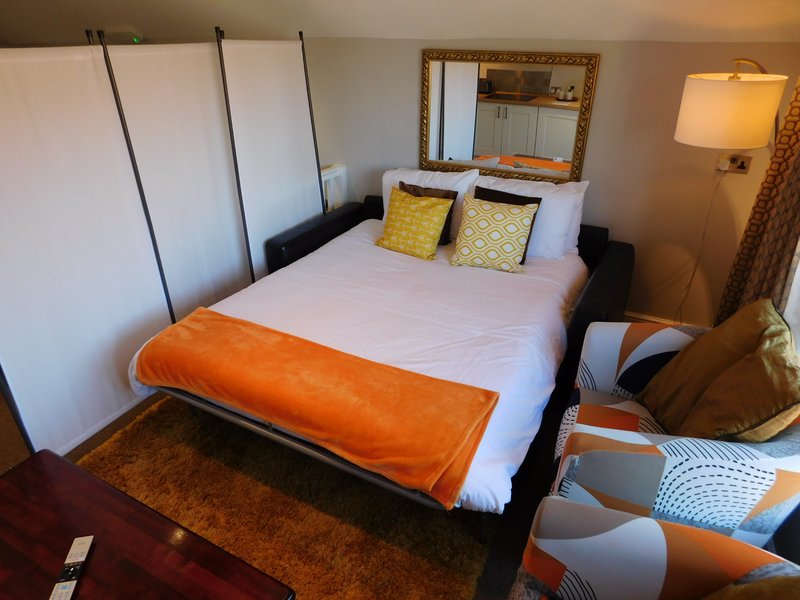 Modern bedroom with double bed, orange throw, yellow cushions, mirror, and bedside lamp