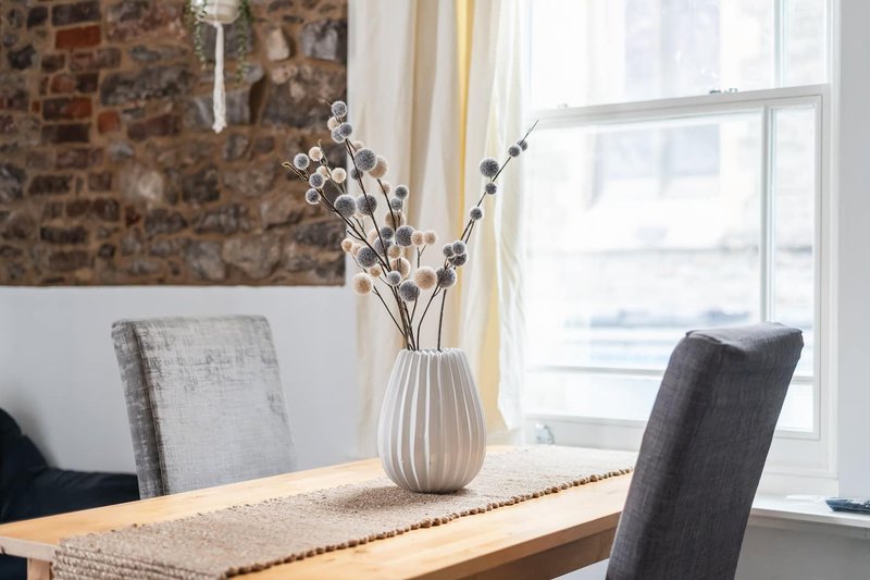 Dining table with chairs, white vase with dried flowers, large window with brick wall