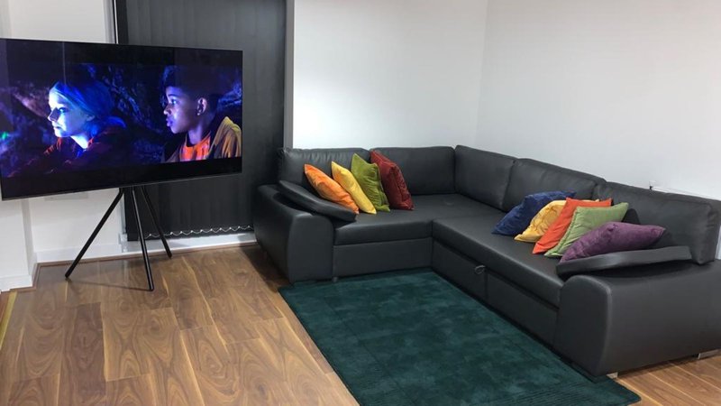 Living room with black corner sofa, colorful cushions, wooden floor, teal rug, and wall-mounted TV