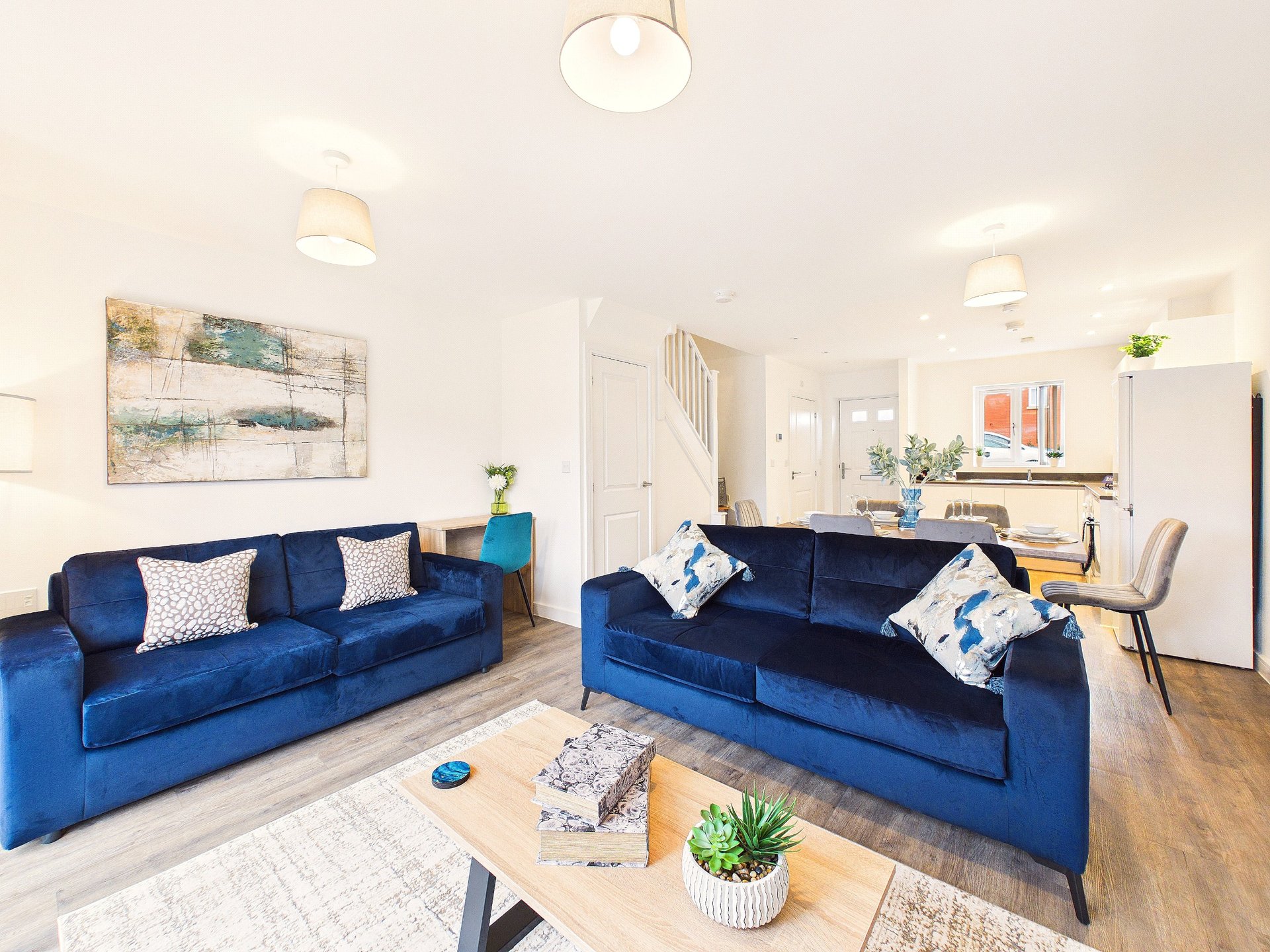 Bright modern living room with two blue sofas, wooden table, pendant lights and open-plan kitchen