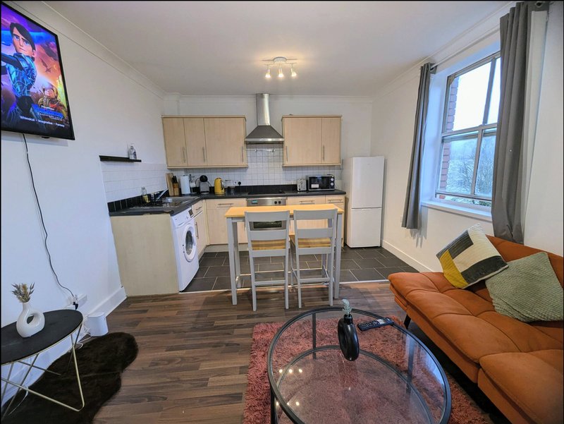 Open-plan kitchen and living area with wooden cabinets, dining table, orange sofa and wall-mounted TV
