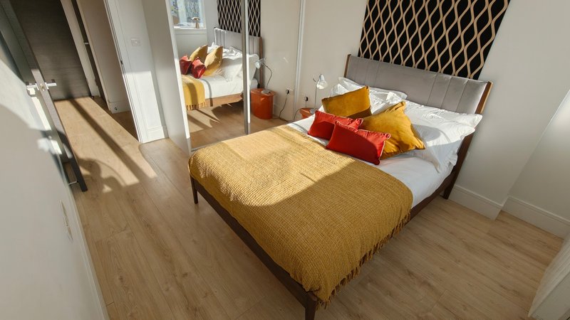 Bright modern bedroom with double bed, mustard bedspread, red and yellow pillows, wooden flooring and ensuite visible