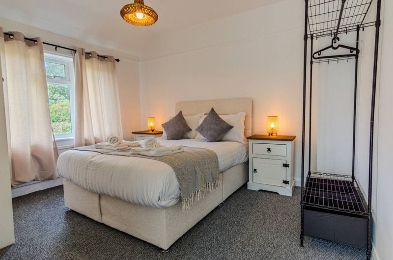 Bright bedroom with double bed, cream bedding, grey cushions, white nightstands with lamps, and garden view through window