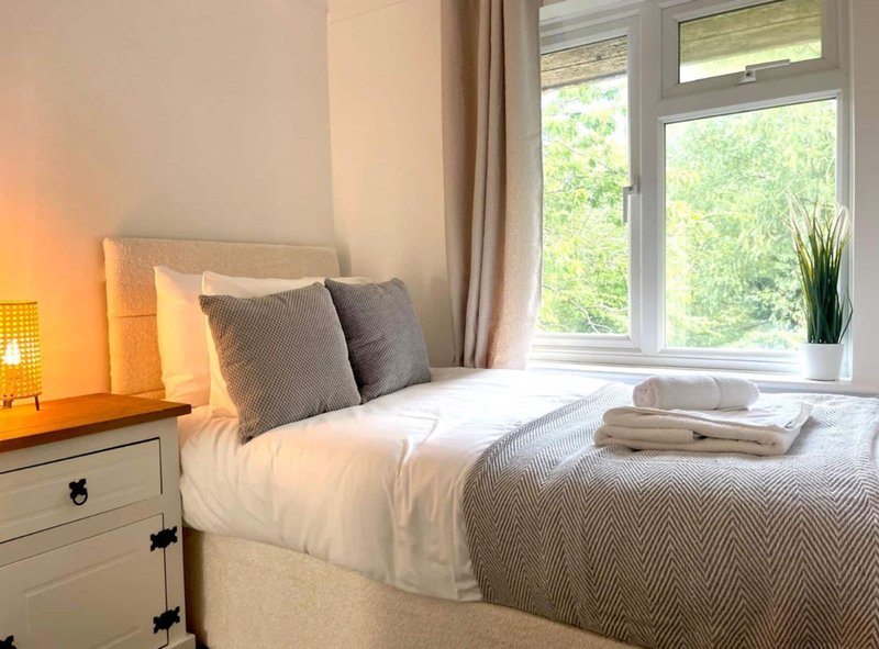 Bright bedroom with single bed, gray cushions, white bedside table, and large window overlooking garden