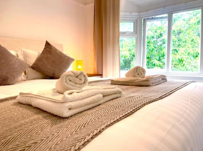 Bedroom with neatly arranged towels on bed, pillows, bedside lamp and large window with garden view