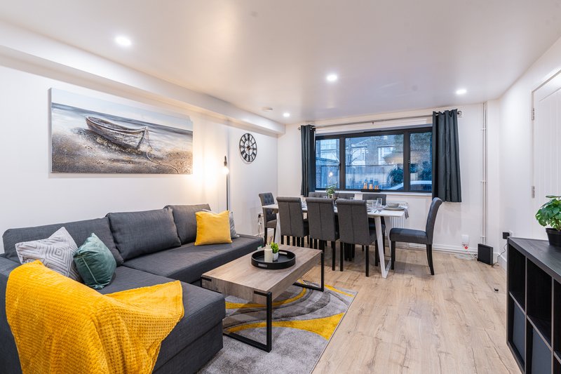 Modern open-plan living room with gray sofa, yellow accent chair, dining table, and large windows