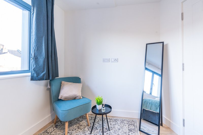 Bright bedroom corner with blue armchair, small side table, standing mirror and window with curtains