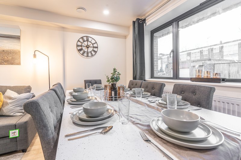 Modern dining area with set table, grey upholstered chairs, wall clock and large windows