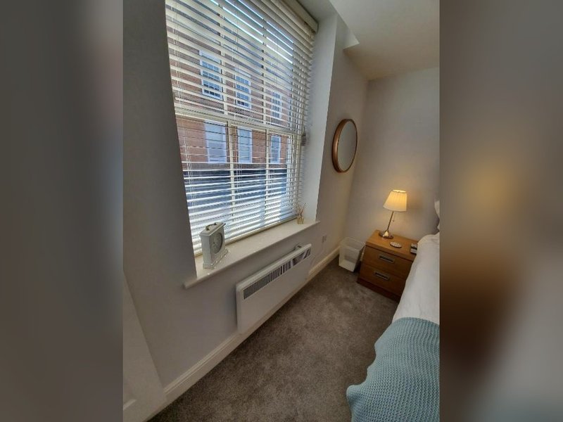 Narrow bedroom with single bed, window with blinds, wooden dresser and bedside lamp