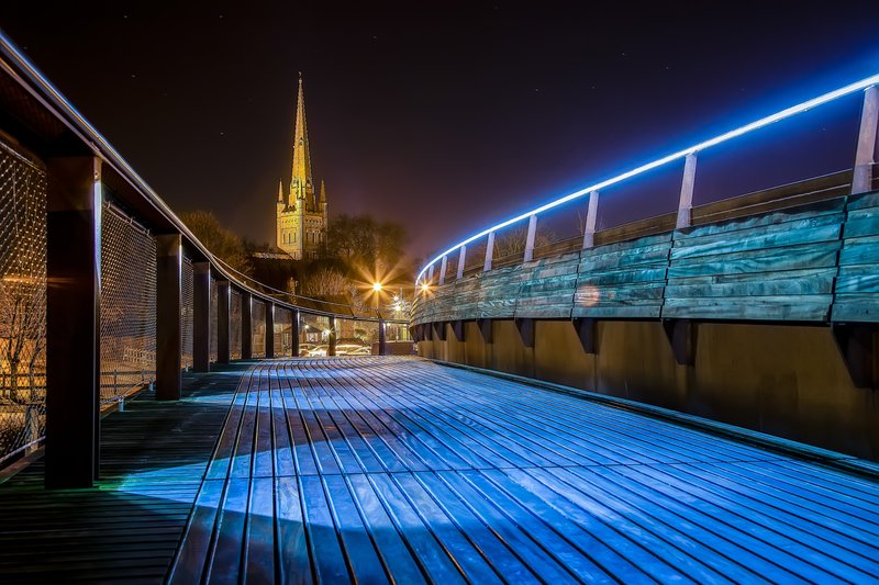 Nighttime view of illuminated wooden deck with blue lighting, church spire visible in background