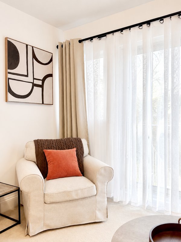 Cream armchair with decorative cushions near window with white curtains and wall art
