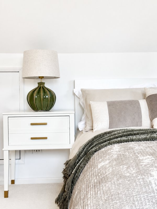 Bedroom nightstand with green lamp, white drawers, and bed with neutral bedding and pillows