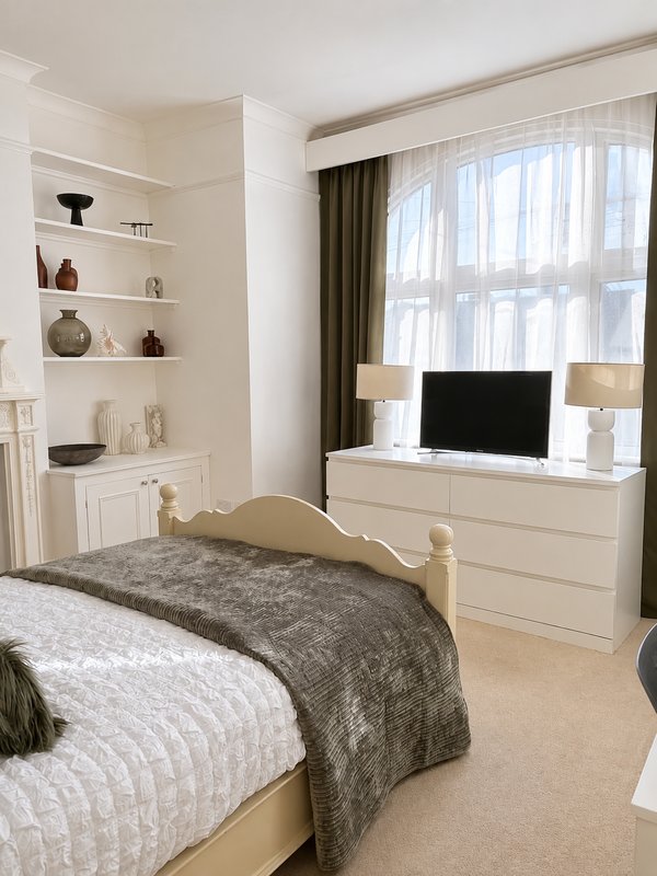 Modern bedroom with double bed, cream furniture, shelving, TV stand and large window with curtains