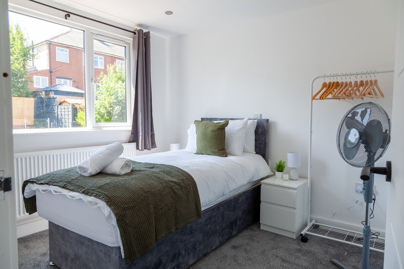 Bedroom with double bed, white bedside table, window with brown curtains, and standing fan