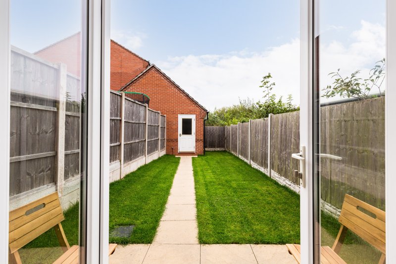 View through patio doors to garden with lawn, pathway, wooden fencing and brick house
