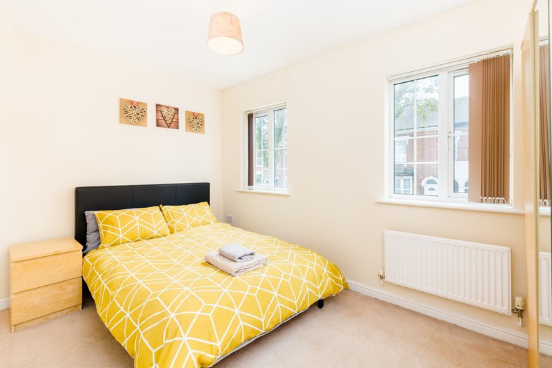 Bright bedroom with yellow patterned duvet, wooden nightstand, and large windows with curtains